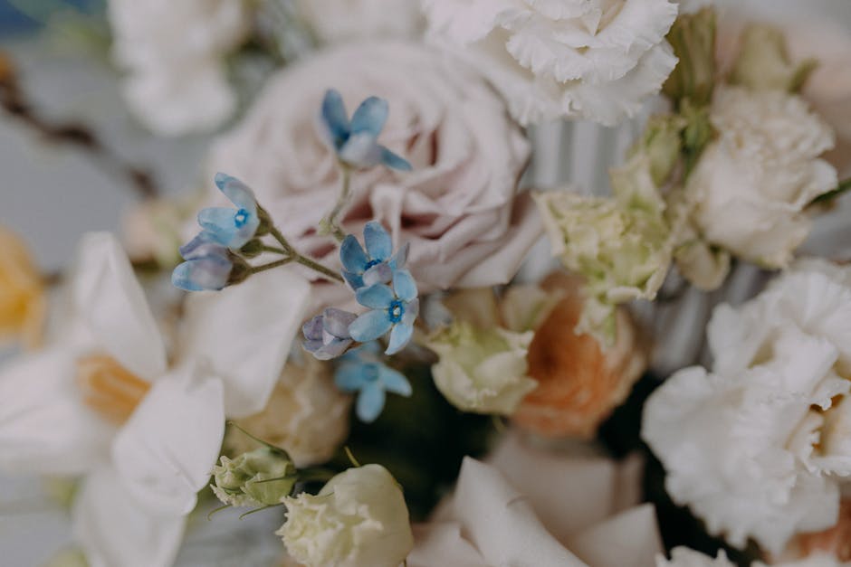 A person’s hand, wearing a light beige long-sleeve shirt, gently holding a cluster of large, vibrant pink dahlias with lush, layered petals. The arrangement is part of a bouquet featuring smaller wh