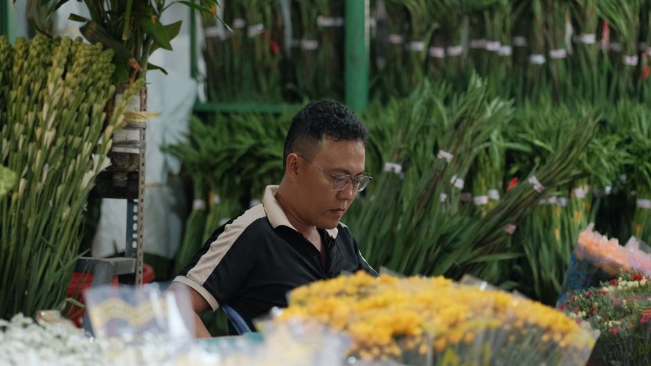 A woman with long dark hair and glasses is holding a bouquet of fresh daisies with white petals and yellow centers, arranged in a clear glass vase. She is wearing a light green short-sleeved shirt and