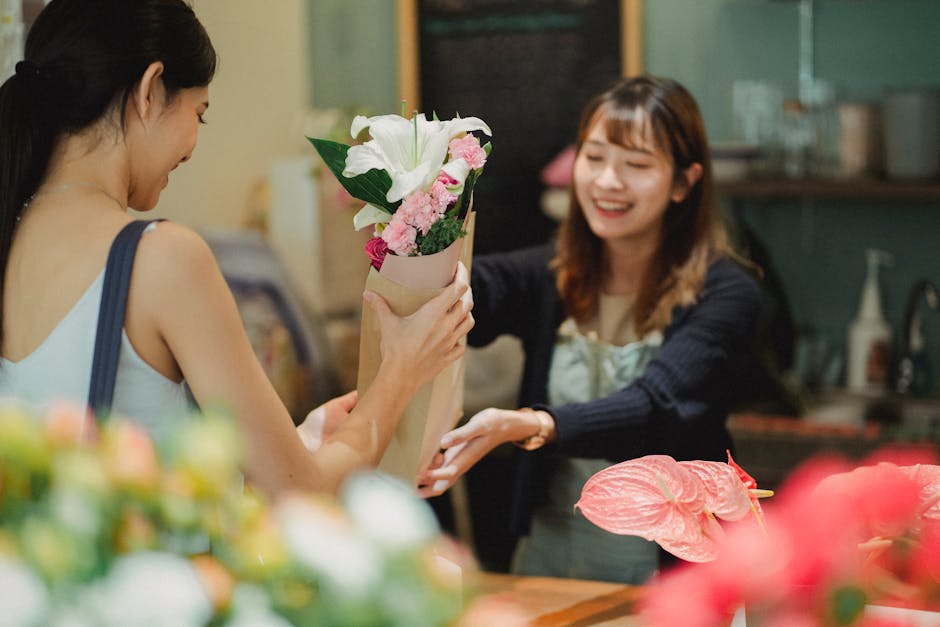 A woman with dark skin, wearing a light blue face mask, black top, and black trousers, is walking down a corridor holding a bouquet of fresh yellow roses wrapped in clear plastic with green foliage, a