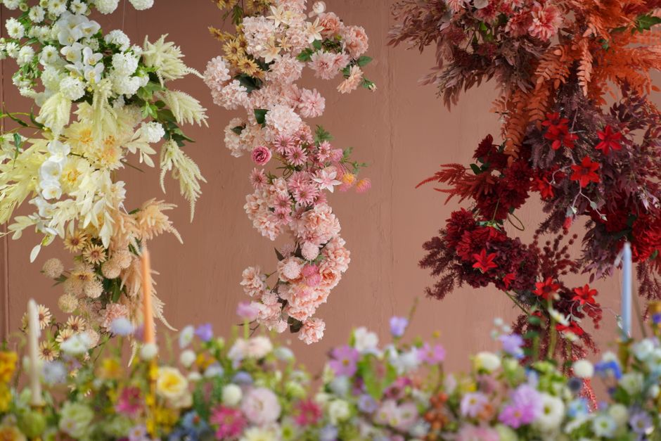 A woman with curly brown hair, dressed in a dark green long-sleeved top, is carefully arranging large artificial peony flowers in soft pink and white colours with textured, ruffled petals. She is usin