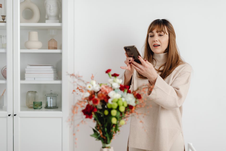 A woman with auburn hair and bangs, dressed in a beige sweater, is holding a vibrant bouquet of fresh flowers in front of her, with a white cabinet filled with decorative vases and books in the backgr