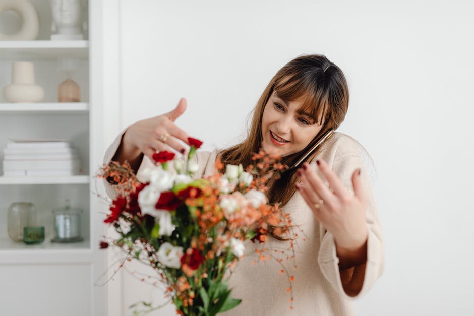 A woman with auburn hair and bangs, dressed in a beige sweater, is holding a vibrant bouquet of fresh flowers in front of her, with a white cabinet filled with decorative vases and books in the backgr